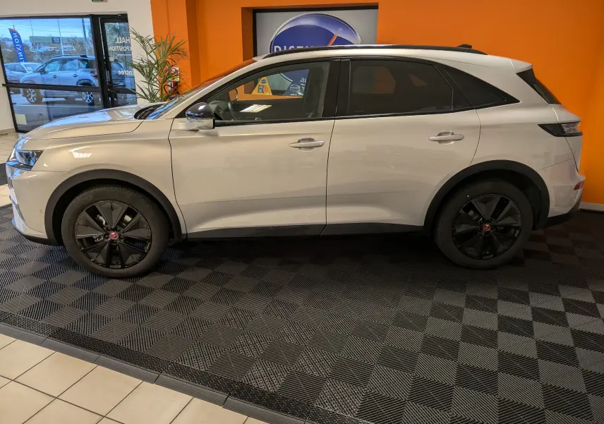 Vue de profil côté gauche d'un DS7 2023 Chrystal Pearl avec jantes noires et barres de toit en showroom.
