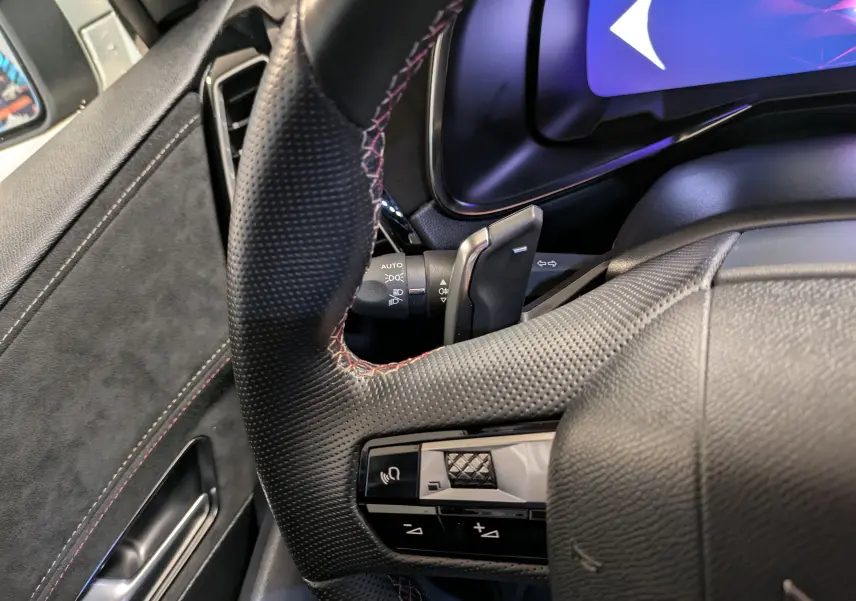 Gros plan sur le volant en cuir perforé noir avec surpiqûres rouges du DS7 2023, tableau de bord numérique en arrière-plan.