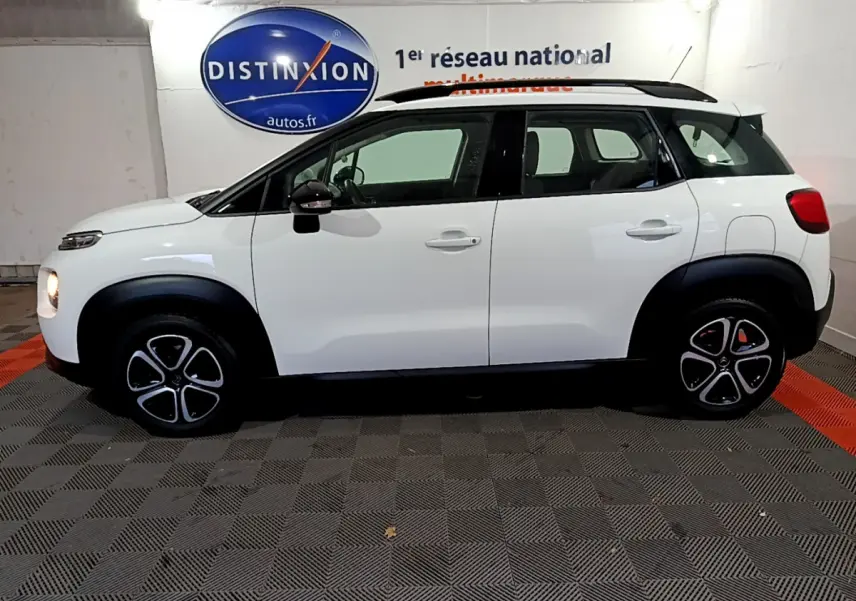 Vue de profil côté gauche d'un Citroën C3 Aircross blanc avec toit noir et jantes distinctives en intérieur showroom.