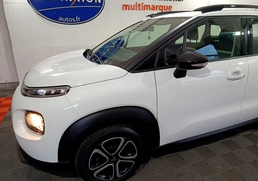Vue de profil avant droit d'une Citroën C3 Aircross blanche avec feux avant allumés et rétroviseurs noirs.