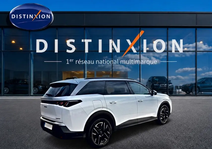 Peugeot 5008 Hybrid blanc en 3/4 arrière droit, avec jantes noires et toit noir, devant un bâtiment vitré Distinxion.
