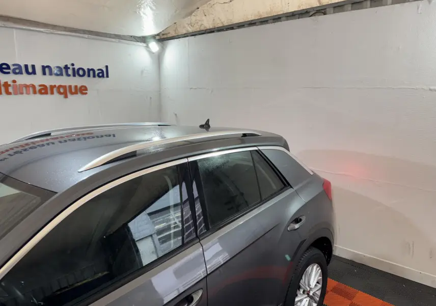 Vue 3/4 arrière droite d'un Volkswagen T-Roc gris foncé avec barres de toit argentées dans un garage.