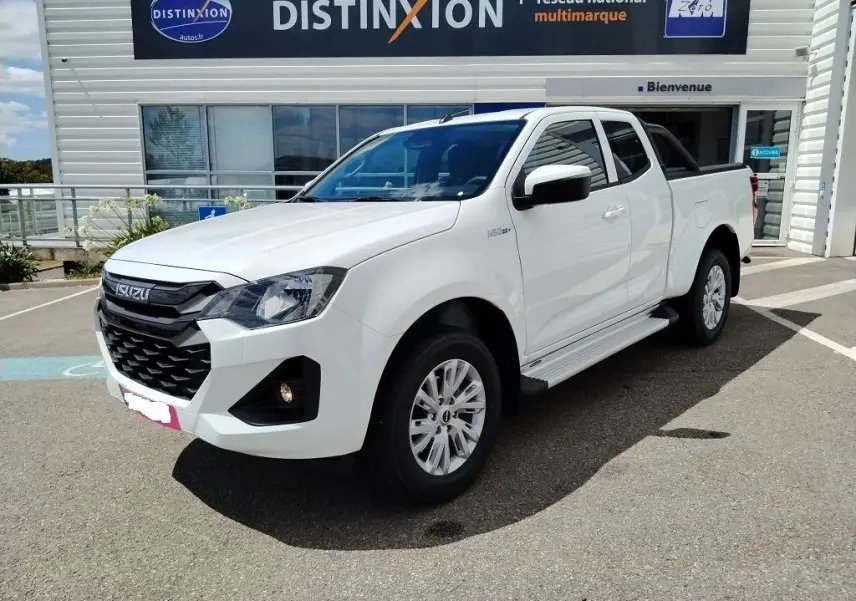 Vue de profil côté droit d'un pick-up Isuzu D-MAX blanc avec arceau noir et couvre-benne roulant, stationné devant un bâtiment industriel.