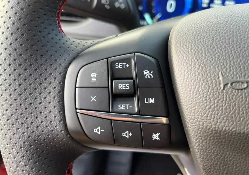 Gros plan sur les commandes de régulateur de vitesse et volume au volant perforé du Ford Kuga 1.5 EcoBoost rouge Lucifer.