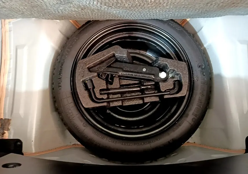 Vue plongeante sur la roue de secours noire et ses outils rangés dans le coffre d'une Citroën C1 blanche.