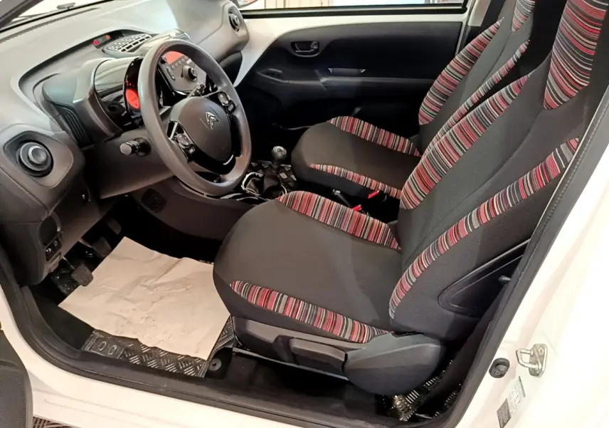Vue intérieure côté conducteur de la Citroën C1 blanche 2020, sièges tissu noir à rayures rouges et volant multifonction.