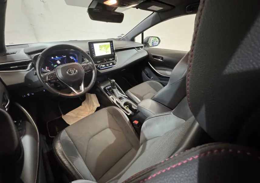 Vue intérieure avant droite de la Toyota Corolla hybride 2021, sièges cuir noir avec surpiqûres rouges et écran tactile central.