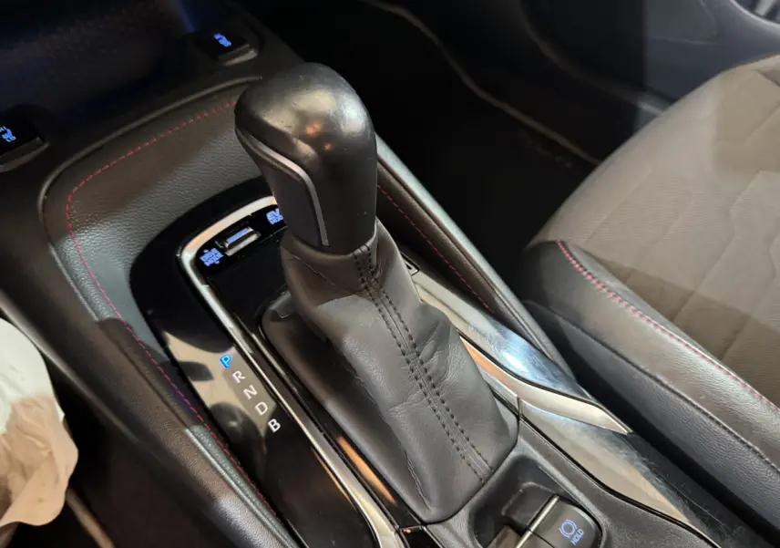 Gros plan sur la console centrale avec levier de vitesse automatique en cuir noir et surpiqûres rouges dans l’habitacle d’une Toyota Corolla gris clair.