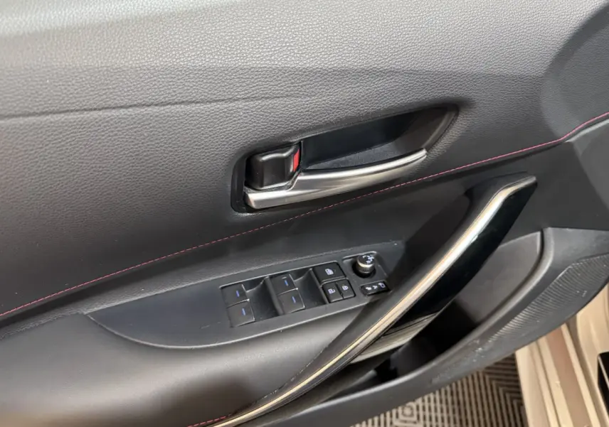 Gros plan sur la poignée intérieure et les commandes de porte côté conducteur d'une Toyota Corolla gris clair, avec surpiqûres rouges.