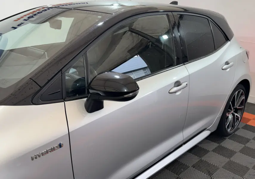 Vue côté gauche d’une Toyota Corolla Hybride gris clair avec toit et rétroviseur noir brillant, badge Hybrid visible.