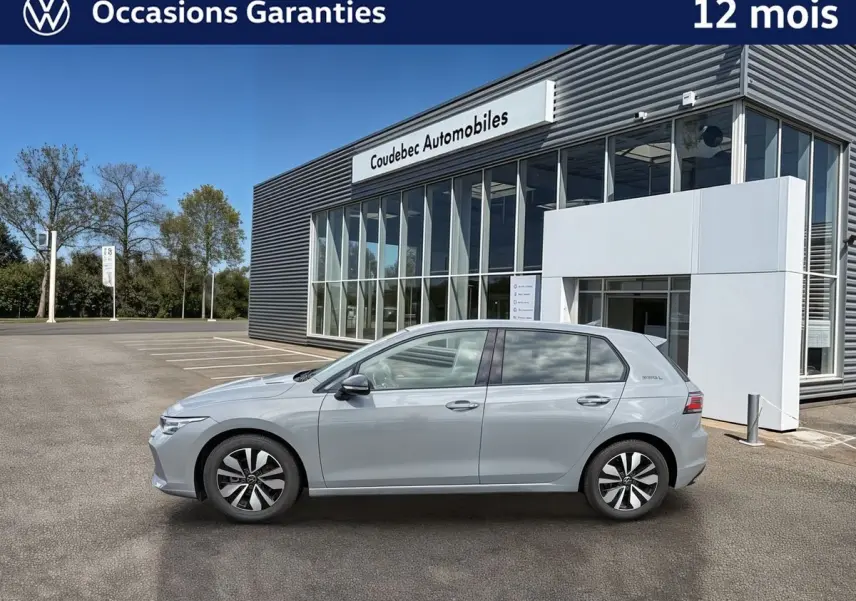 Profil côté gauche d'une Volkswagen Golf 1.5 eTSI 150 DSG7 grise, garée devant un bâtiment moderne de concession.