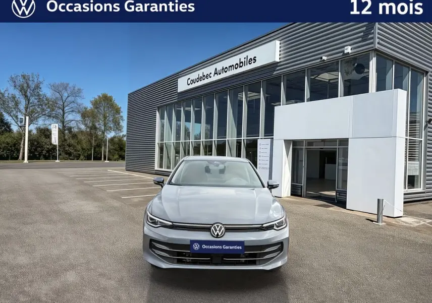 Vue frontale d'une Volkswagen Golf 1.5 eTSI grise stationnée devant un bâtiment moderne de concession automobile.