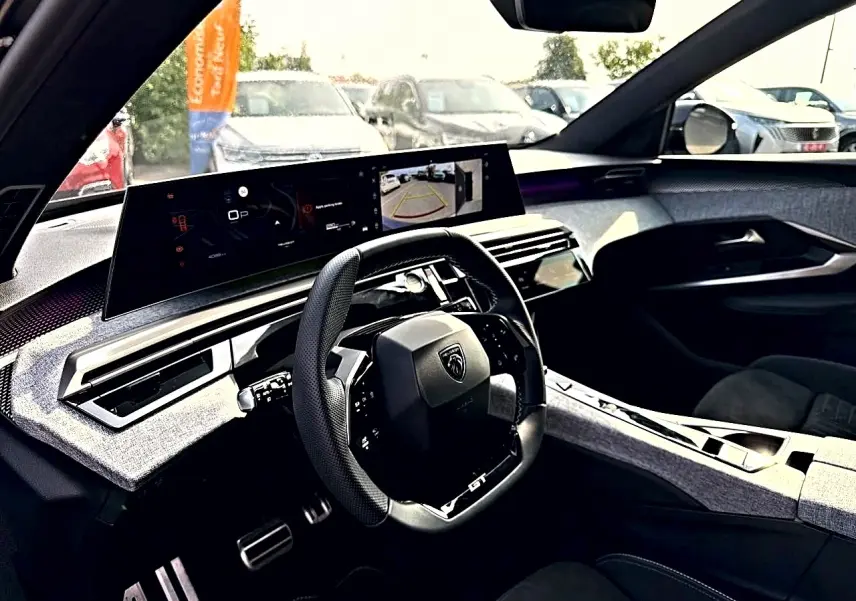 Vue intérieure côté conducteur du Peugeot 5008 Hybrid 145 GT e-dcs6 2025, tableau de bord moderne avec écran large et volant cuir noir.