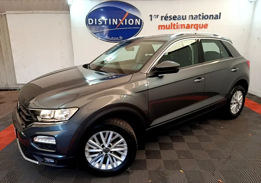 Volkswagen T-Roc gris foncé vue 3/4 avant droit, avec jantes alliage et barres de toit argentées visibles.