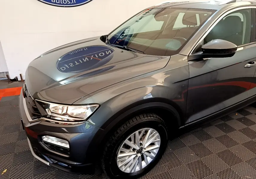Vue 3/4 avant droit d'un Volkswagen T-Roc gris foncé avec jantes alliage et phares LED visibles en intérieur showroom.