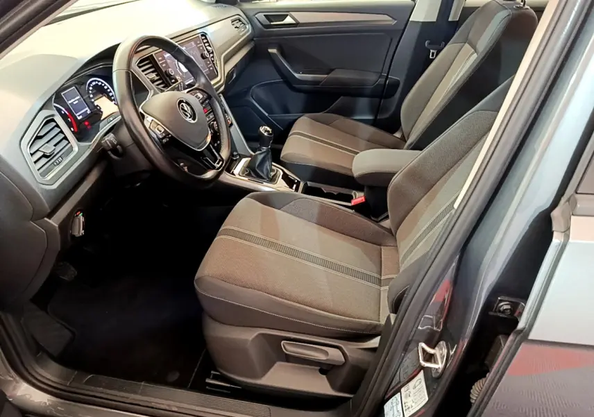 Vue intérieure côté conducteur du Volkswagen T-Roc gris foncé, montrant le volant cuir, tableau de bord digital et sièges tissu gris.