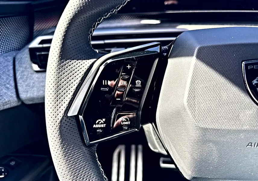 Gros plan sur le volant cuir perforé noir du Peugeot 5008 Hybrid, avec commandes multifonctions visibles à gauche.