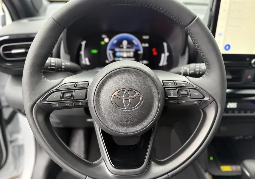 Vue rapprochée du volant noir de la Toyota Yaris Cross 130 CH Design 2025 avec commandes intégrées et tableau de bord numérique flou.