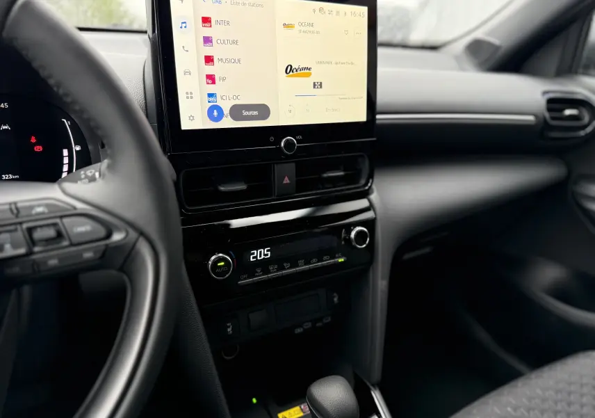 Vue intérieure centrée sur la console centrale et l'écran tactile du Toyota Yaris Cross blanc pur 2025, avec volant partiel visible.