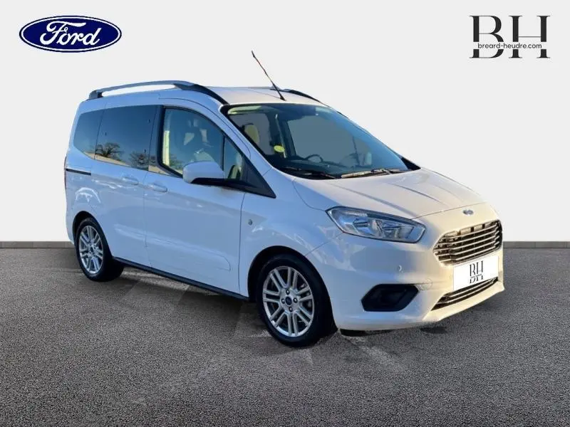 Ford Tourneo Courier blanc glacier en vue 3/4 avant droit, avec jantes alu et barres de toit visibles.