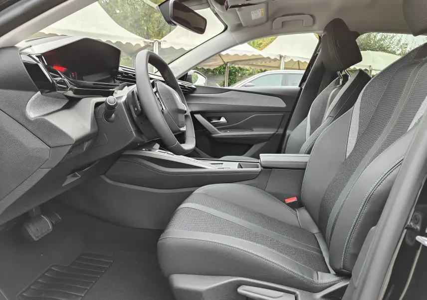Vue intérieure côté conducteur de la Peugeot 308 PureTech 130 Allure Pack 2023, sièges noirs avec surpiqûres bleues et tableau de bord moderne.