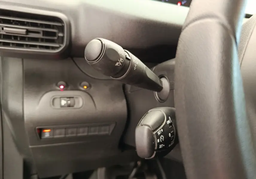 Gros plan sur la commande des feux et régulateur de vitesse à gauche du volant dans l’habitacle noir du Peugeot Partner blanc.