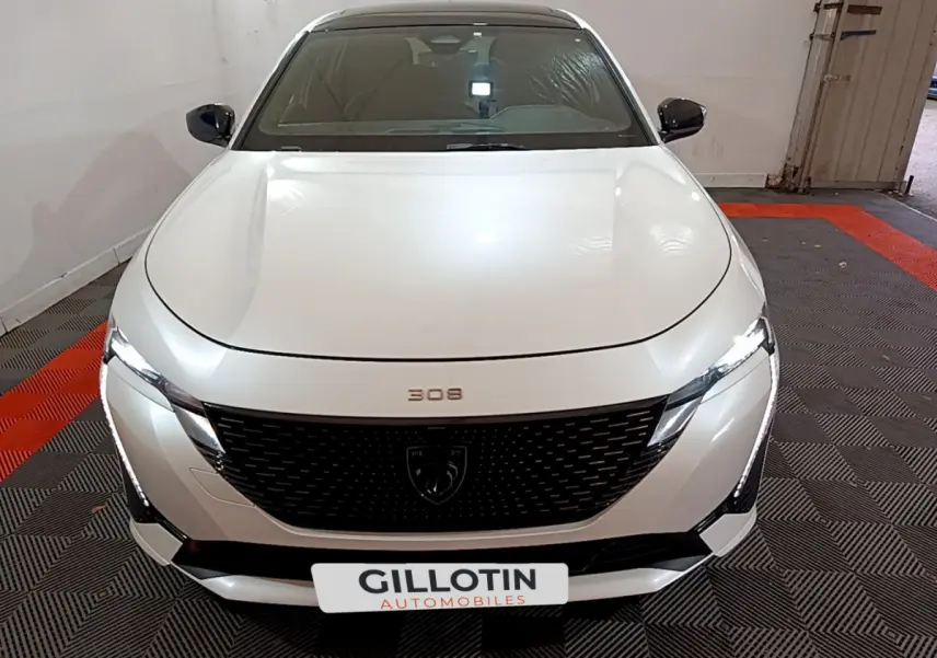 Vue frontale d'une Peugeot 308 PHEV 180 E-EAT8 GT blanche avec calandre noire et phares LED allumés dans un garage.