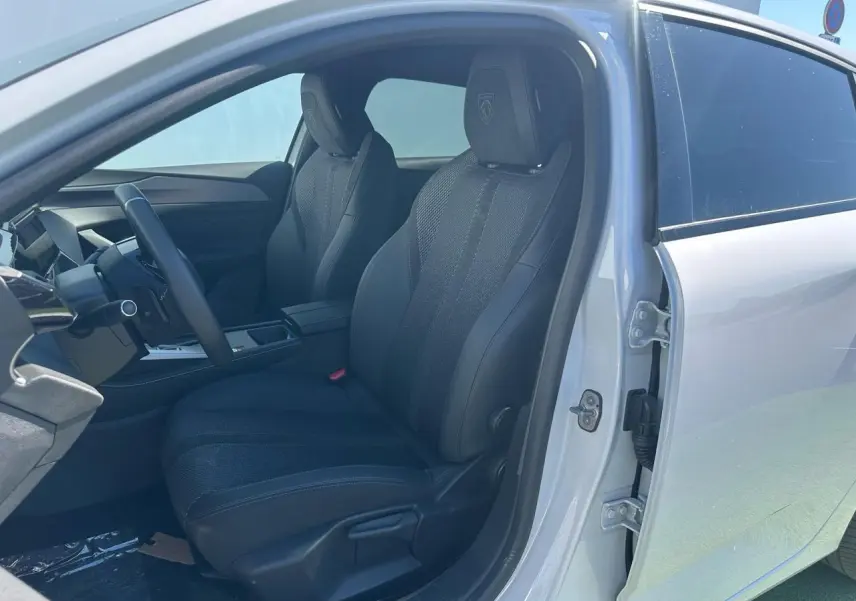 Vue intérieure côté conducteur de la Peugeot 308 blanche Okenite, sièges noirs avec logo Peugeot brodé sur appuie-tête.