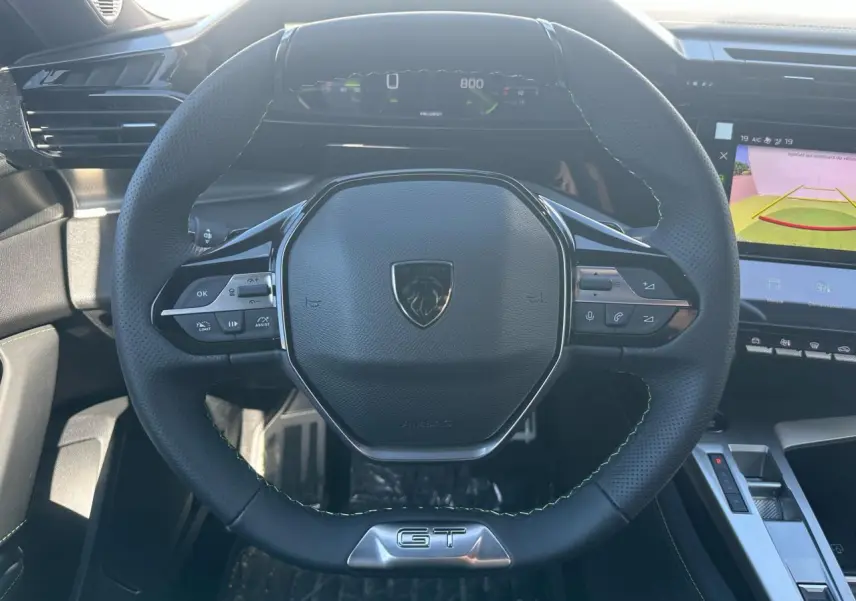 Volant cuir noir GT de Peugeot 308 2025 vu de face, avec commandes intégrées et combiné numérique en arrière-plan.