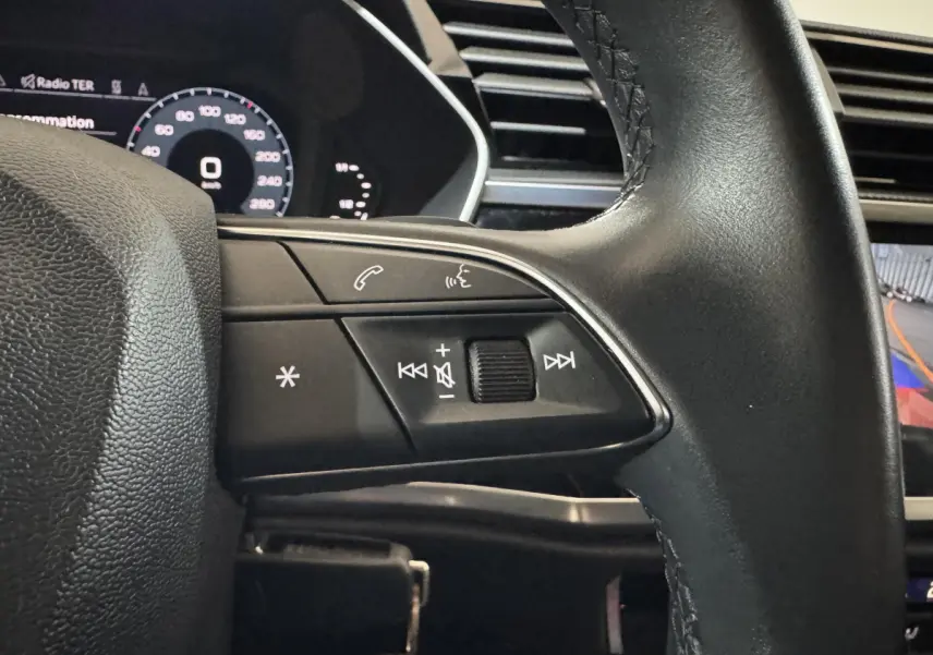 Gros plan sur le volant noir de l'Audi Q3 2022, avec commandes audio et affichage numérique partiel en arrière-plan.