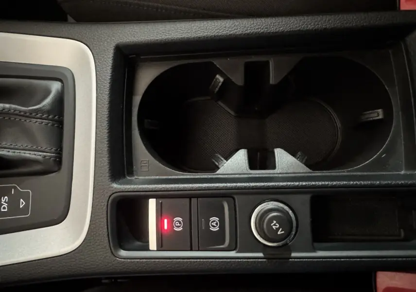 Gros plan sur la console centrale noire de l'Audi Q3 2022, avec porte-gobelets, bouton frein à main électrique et prise 12V.