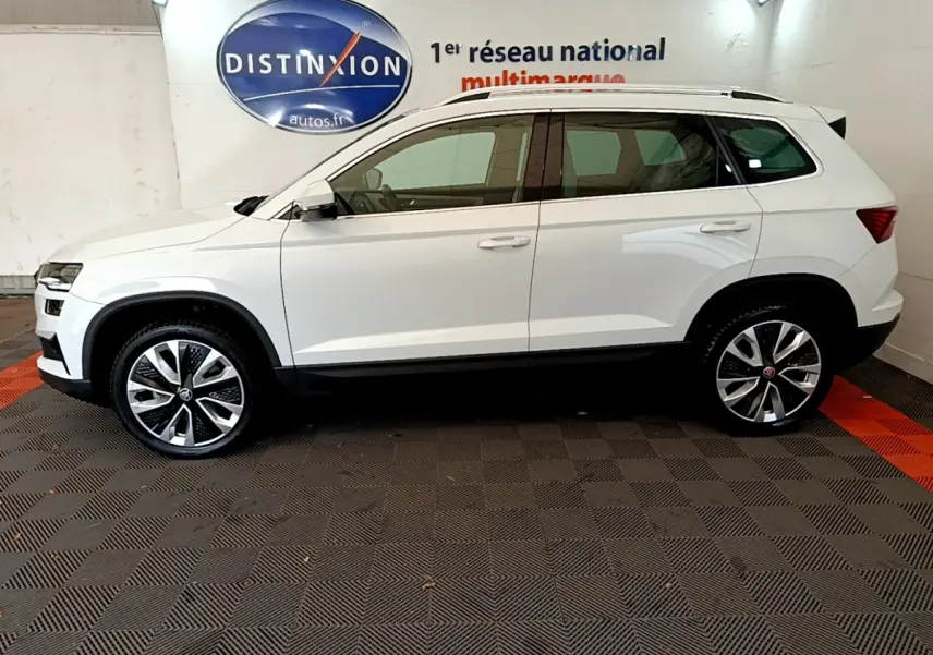 Vue latérale droite d'un Skoda Karoq blanc 2023 avec jantes alliage et rails de toit chromés en intérieur showroom.
