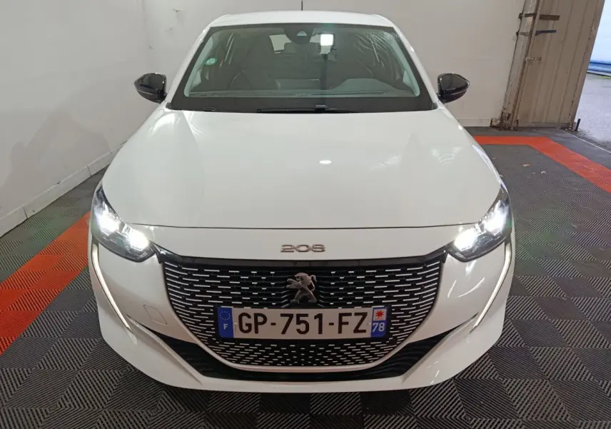 Vue frontale d'une Peugeot 208 électrique blanche avec phares LED allumés et calandre noire distinctive.
