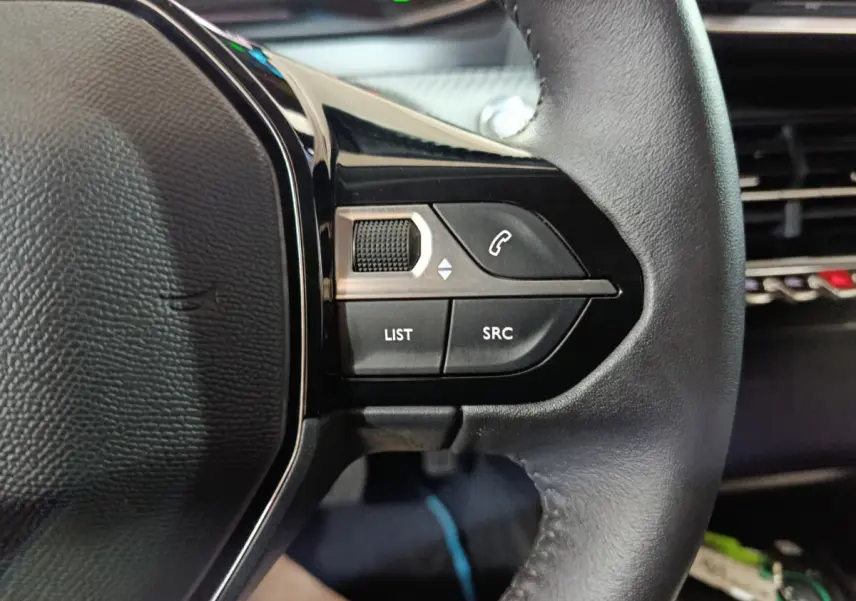 Gros plan sur les commandes gauche du volant cuir d'une Peugeot 208 électrique blanche, avec boutons multifonctions et molette.