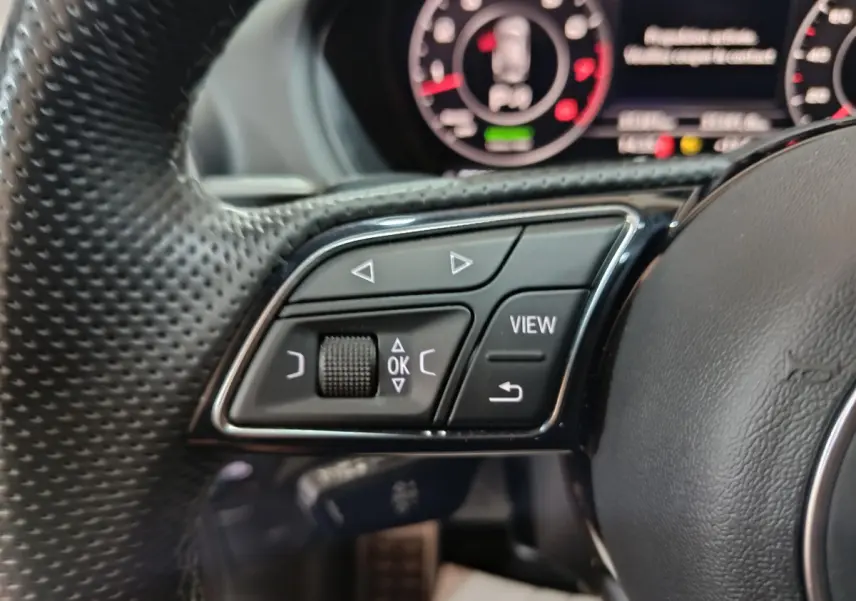 Gros plan sur les commandes gauche du volant cuir perforé d'un Audi Q2 noir, avec tableau de bord numérique en arrière-plan.