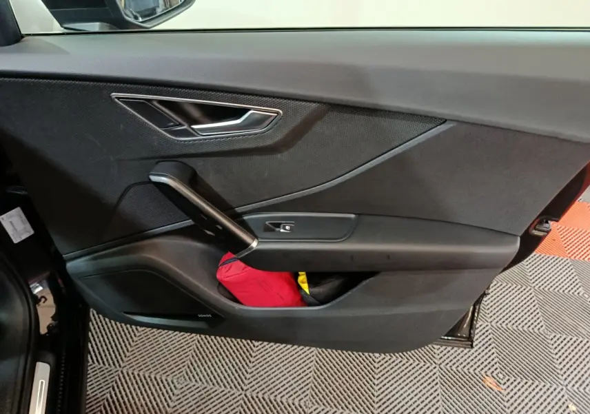 Porte intérieure côté gauche noire de l'Audi Q2 35 TFSI 2022 avec poignée argentée et rangement contenant un objet rouge.