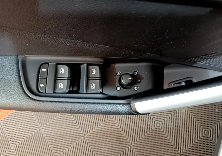 Gros plan sur les commandes de lève-vitres et réglage des rétroviseurs côté conducteur d’un Audi Q2 gris clair, intérieur noir.