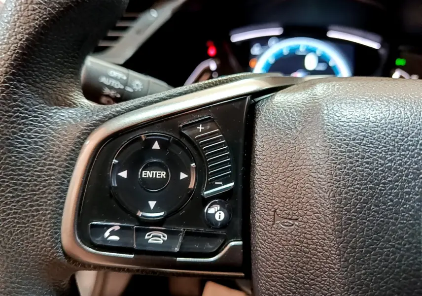 Gros plan sur les commandes gauche du volant cuir de la Honda Civic 1.0 i-VTEC Exclusive 2020, avec tableau de bord lumineux en arrière-plan.