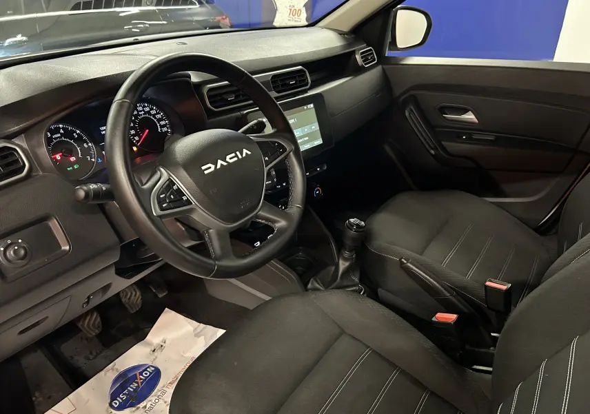 Vue intérieure côté conducteur du Dacia Duster 2024, tableau de bord noir avec volant marqué DACIA et sièges tissu noirs.
