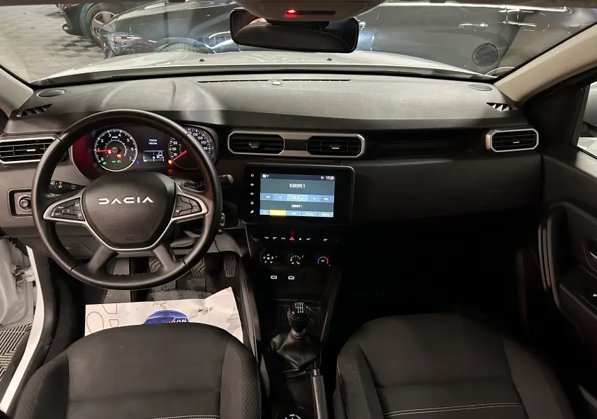 Vue intérieure frontale du Dacia Duster blanc 2024, montrant le tableau de bord, le volant multifonction et l'écran tactile central.