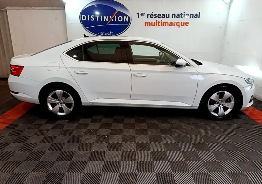 Vue de profil côté gauche d'une Skoda Superb blanche 2021 dans un showroom avec sol à damier noir et rouge.