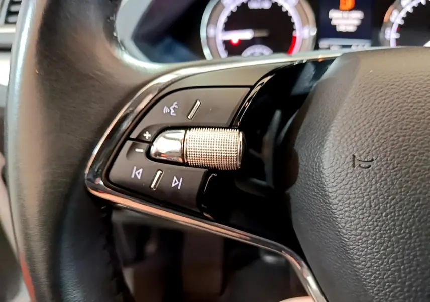 Gros plan sur les commandes gauche du volant cuir noir de la Skoda Superb blanche, avec tableau de bord numérique en arrière-plan.