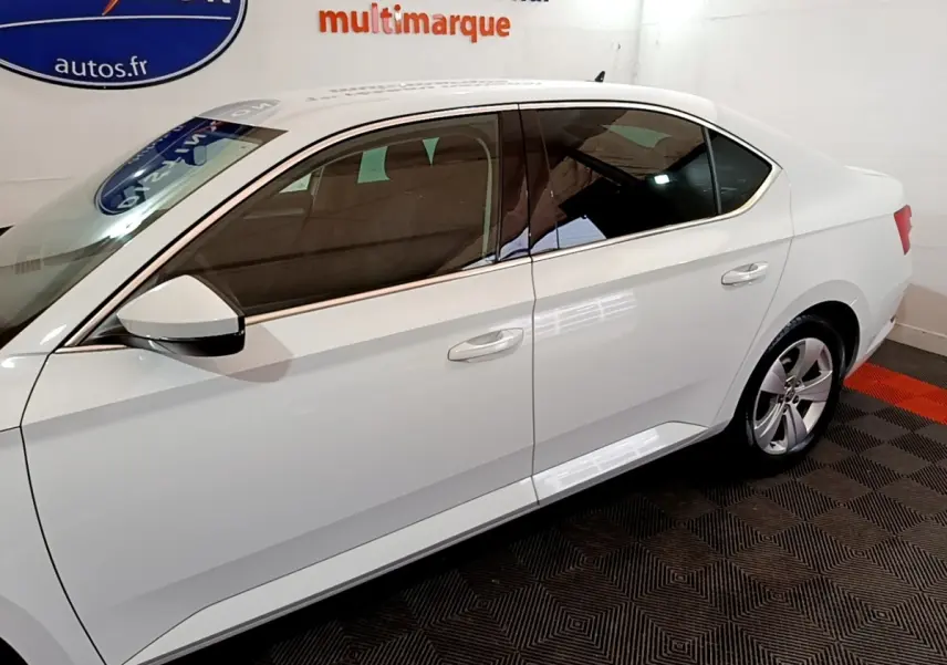 Vue latérale droite d'une Skoda Superb blanche 2021 dans un showroom avec jantes alliage et vitres teintées.