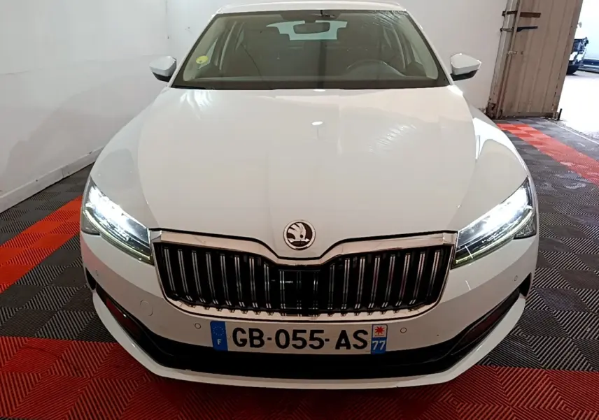 Vue avant d'une Skoda Superb blanche 2021 avec phares LED allumés et calandre chromée dans un garage.