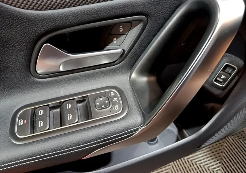 Gros plan sur la poignée intérieure et les commandes de vitres côté gauche d'une Mercedes CLA 180 d Business Line gris clair.