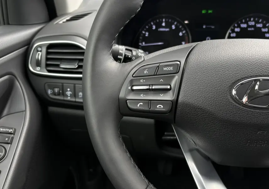Gros plan sur le volant cuir gris foncé de la Hyundai i30 2025, avec commandes audio et tableau de bord en arrière-plan.