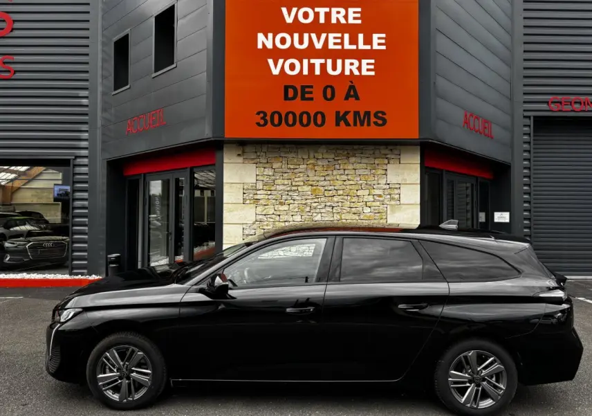 Profil latéral gauche d'une Peugeot 308 noire 2024 garée devant un bâtiment avec panneau orange "Votre nouvelle voiture"