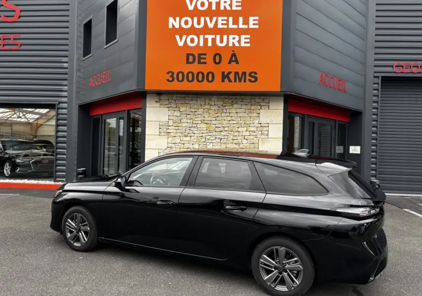 Peugeot 308 PureTech 130 noire vue de profil côté gauche, stationnée devant un bâtiment avec panneau orange.