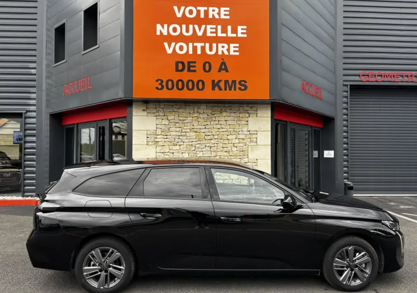 Profil côté gauche d'une Peugeot 308 noire 2024 garée devant un bâtiment avec panneau orange "Votre nouvelle voiture".