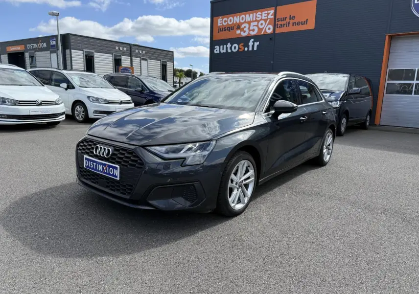 Audi A3 Sportback gris Manhattan métallisé vue 3/4 avant droit sur parking avec enseigne orange en arrière-plan